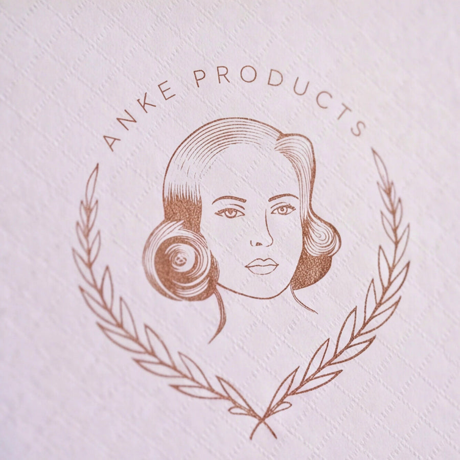 A close-up of a textured white paper surface featuring a circular embossed logo with the text 'ANKE PRODUCTS' in brown around a stylized woman's face and laurel wreath.