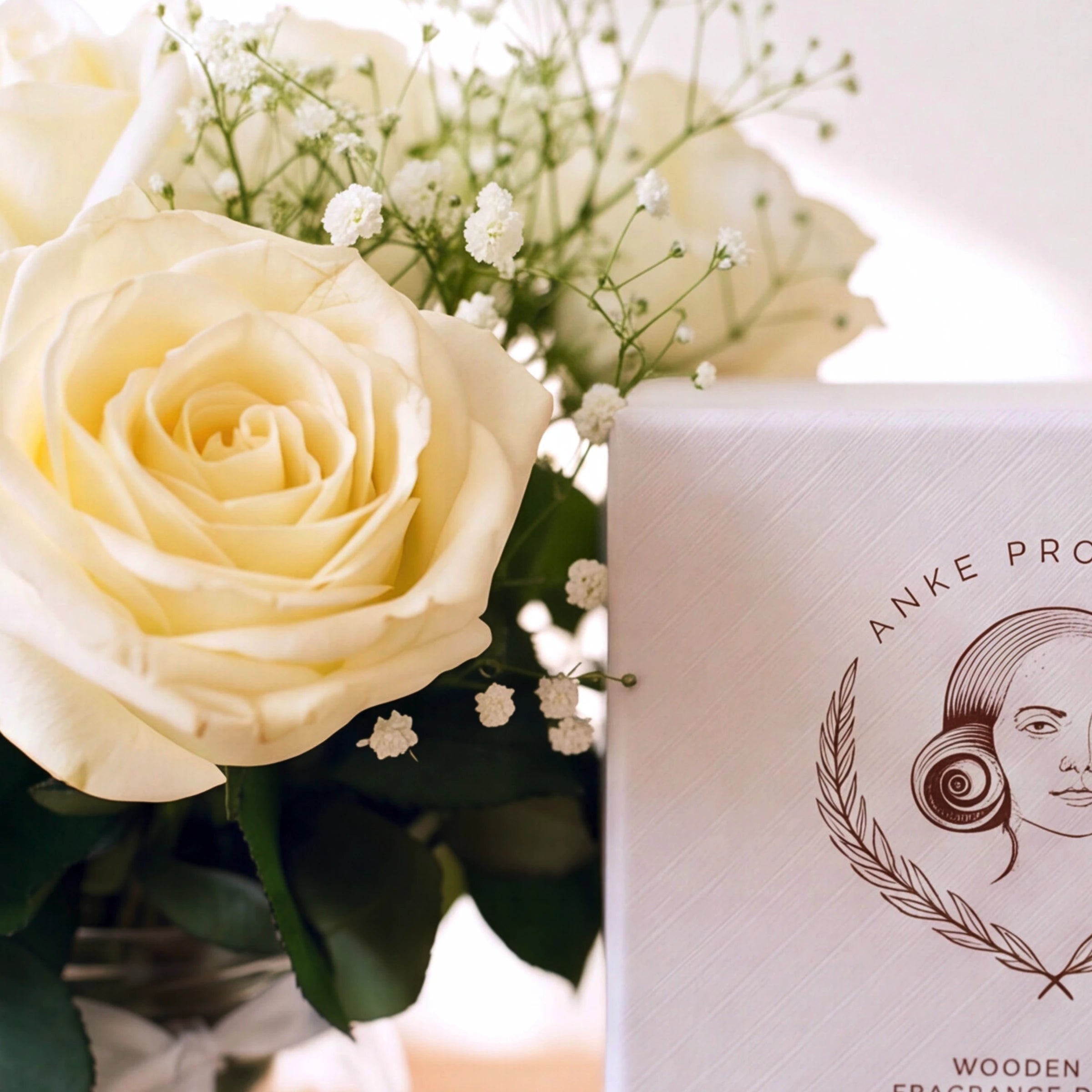 A close-up of a white rose arrangement with small white baby's breath beside a white product box printed with the brand name Anke Pro and a rose illustration. The box text partially reads 'Wooden Fragrance.
