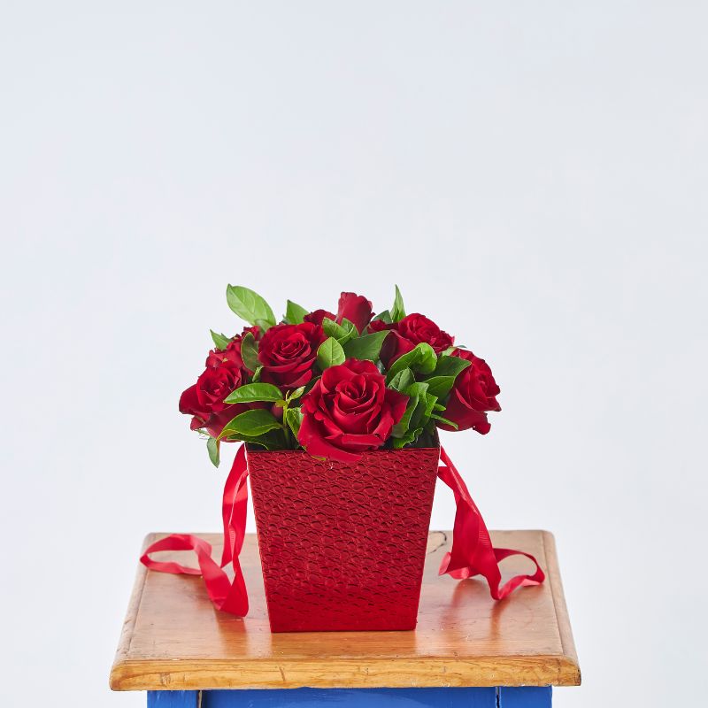 Red Rose Flower Box with ten deep red roses and green foliage in a textured red container with ribbon, displayed on a wooden table.
