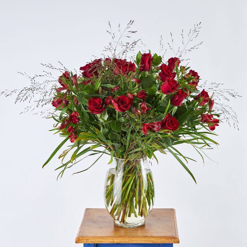 Red Rose of Romance bouquet with two dozen long-stemmed red roses, lush greenery, and filler flowers in a clear glass vase on a wooden table.
