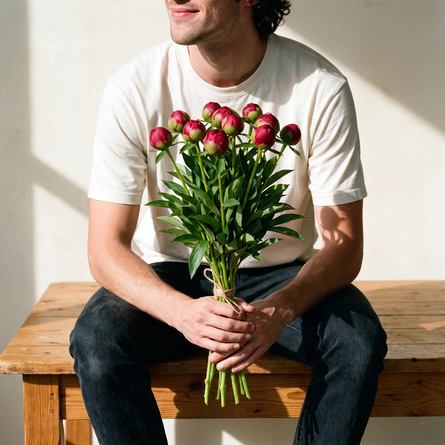 Man seated on a pine table holding a bunch of red peony buds