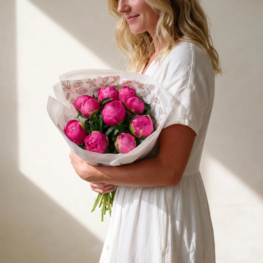 Blonde woman in a white dress walking past a sunlit cream wall with a magenta peony bouquet in the crook of her arm, Fabulous Flowers South Africa