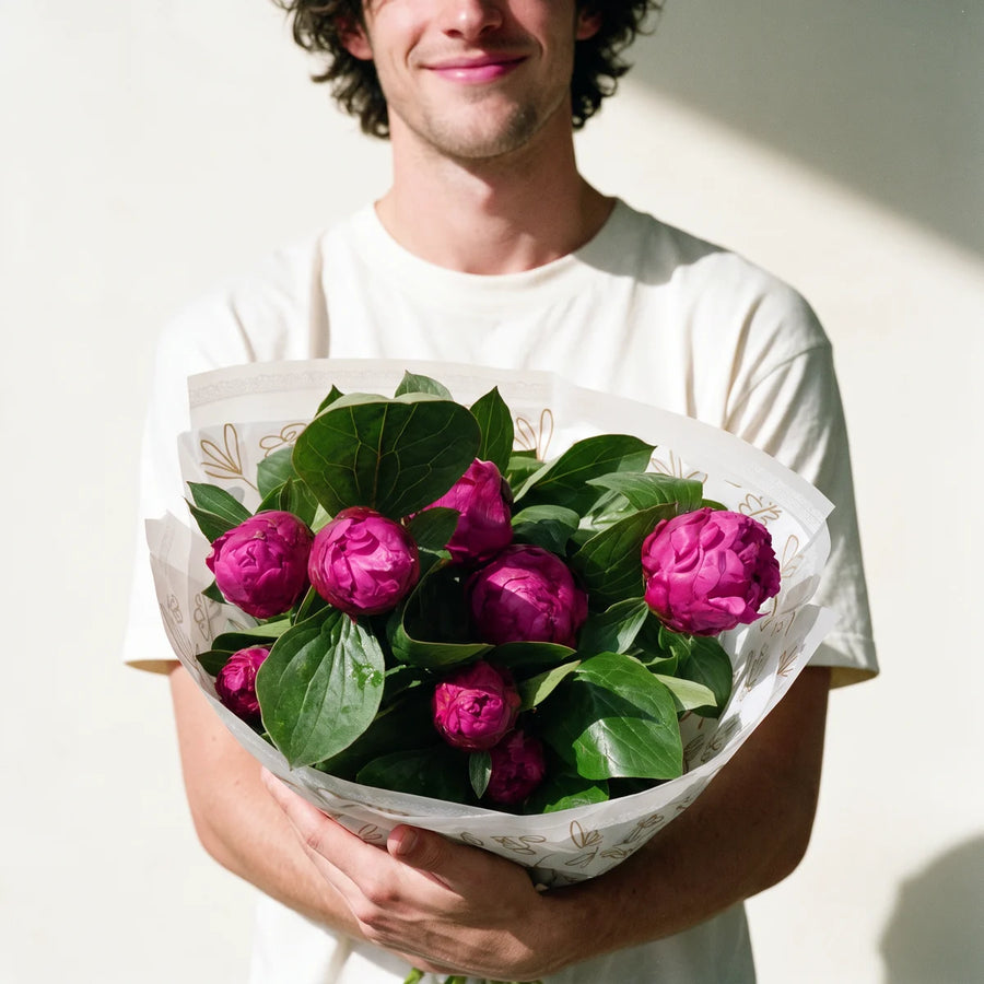 Smiling man with curly dark hair in a white t-shirt holding out a bouquet of ten magenta peonies towards the camera, Fabulous Flowers South Africa