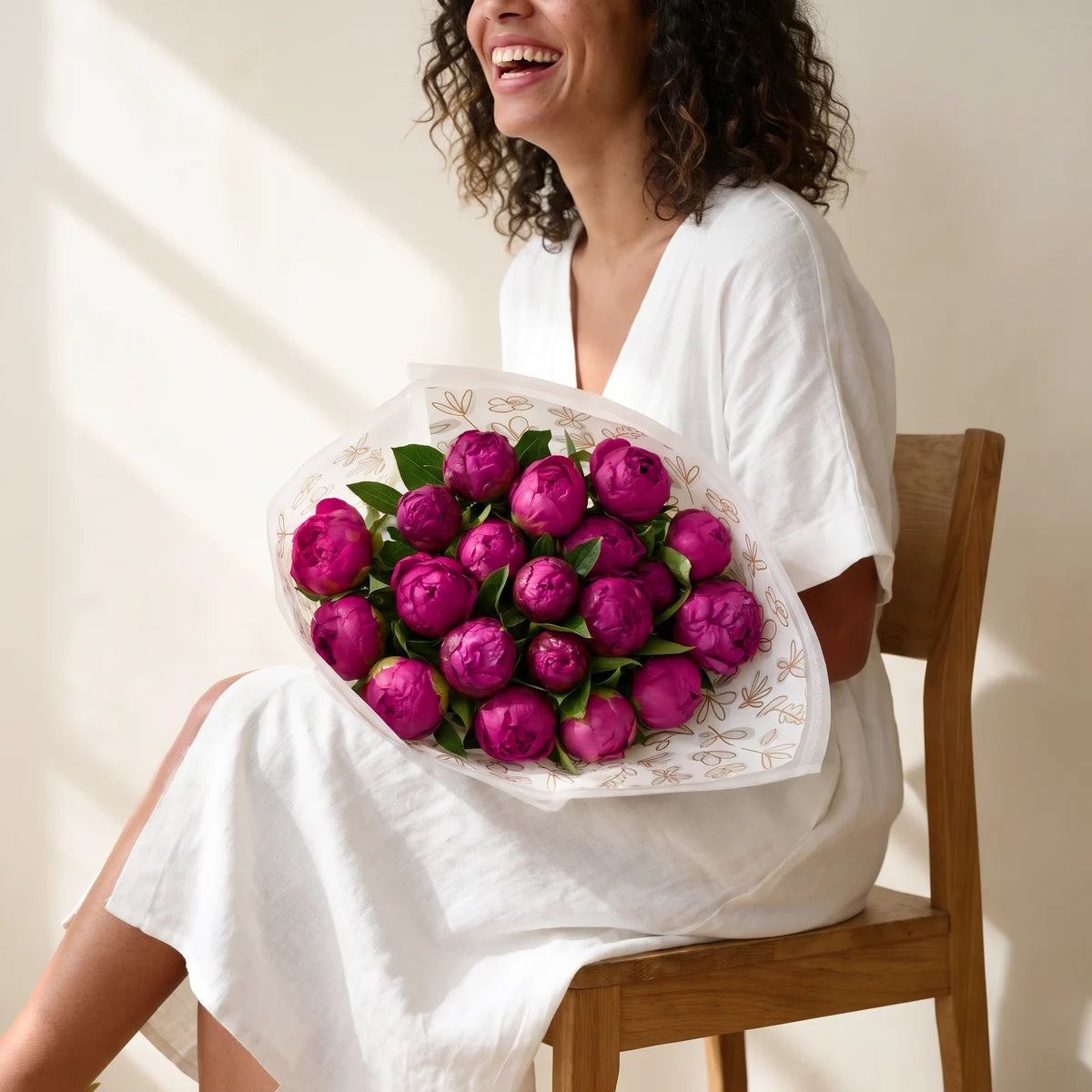 Laughing woman with curly dark hair in a white dress sitting on a wooden chair with a large magenta peony bouquet on her lap, Fabulous Flowers South Africa