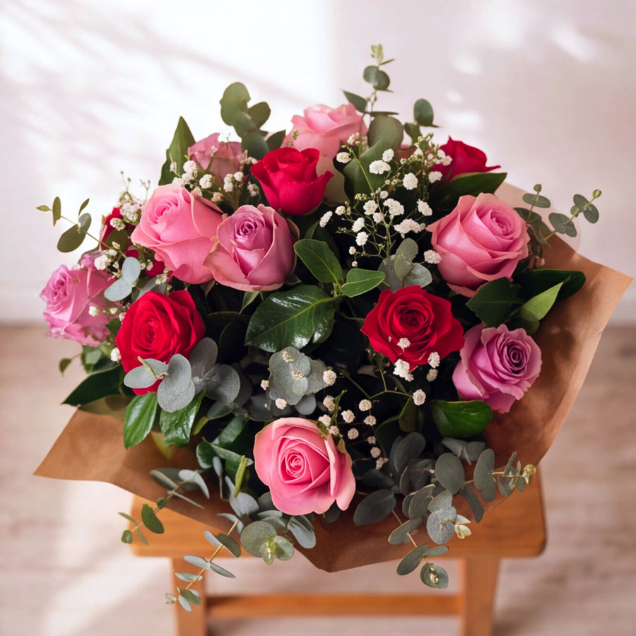 A vibrant floral bouquet featuring pink and red roses, green eucalyptus leaves, and small white baby's breath, wrapped in brown paper and displayed on a wooden stool against a softly lit background.