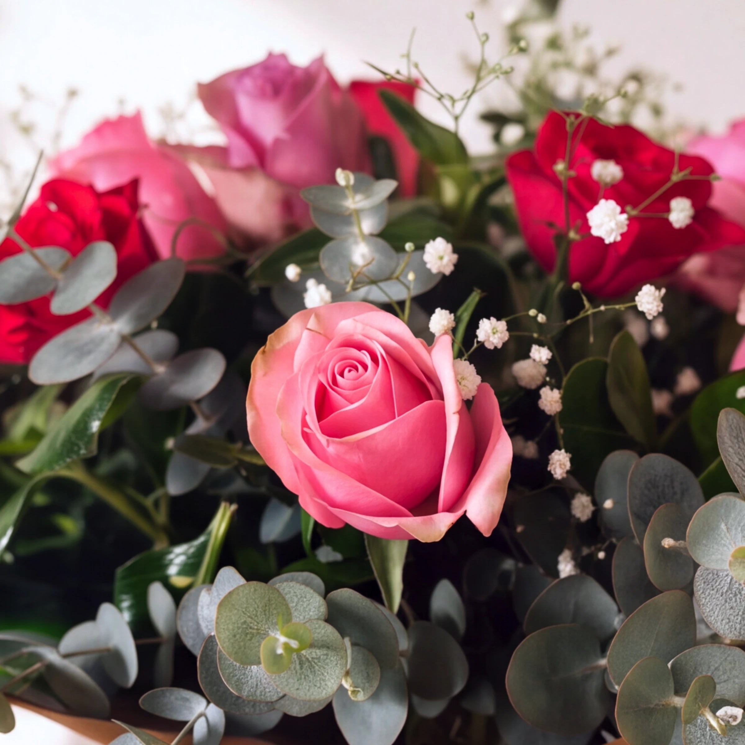 A close-up floral arrangement featuring a prominent pink rose in the foreground, surrounded by red and pink roses, green eucalyptus leaves, and small white baby's breath flowers.