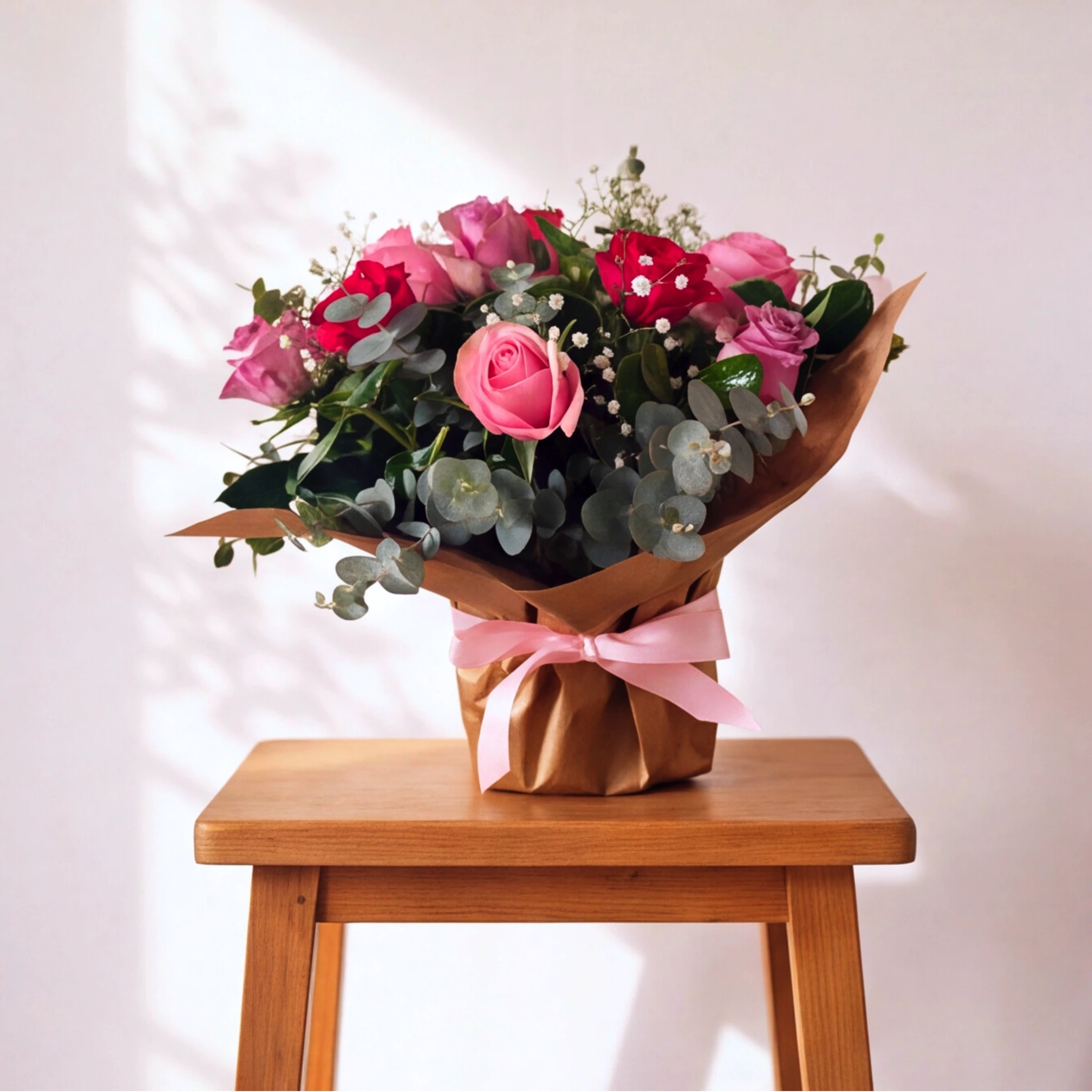 A bouquet of pink and red roses with green eucalyptus leaves and small white filler flowers, wrapped in brown paper with a pink ribbon, displayed on a wooden stool against a light wall with soft shadows.