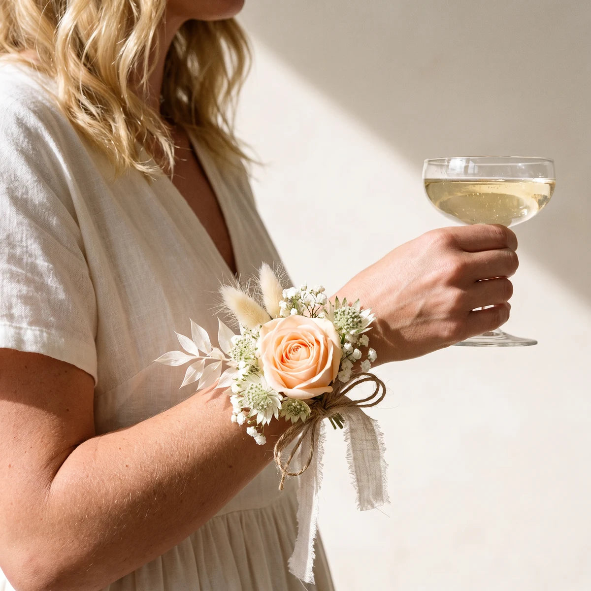 Woman in a cream linen dress holding a coupe of champagne and wearing a rustic peach rose corsage, Fabulous Flowers South Africa