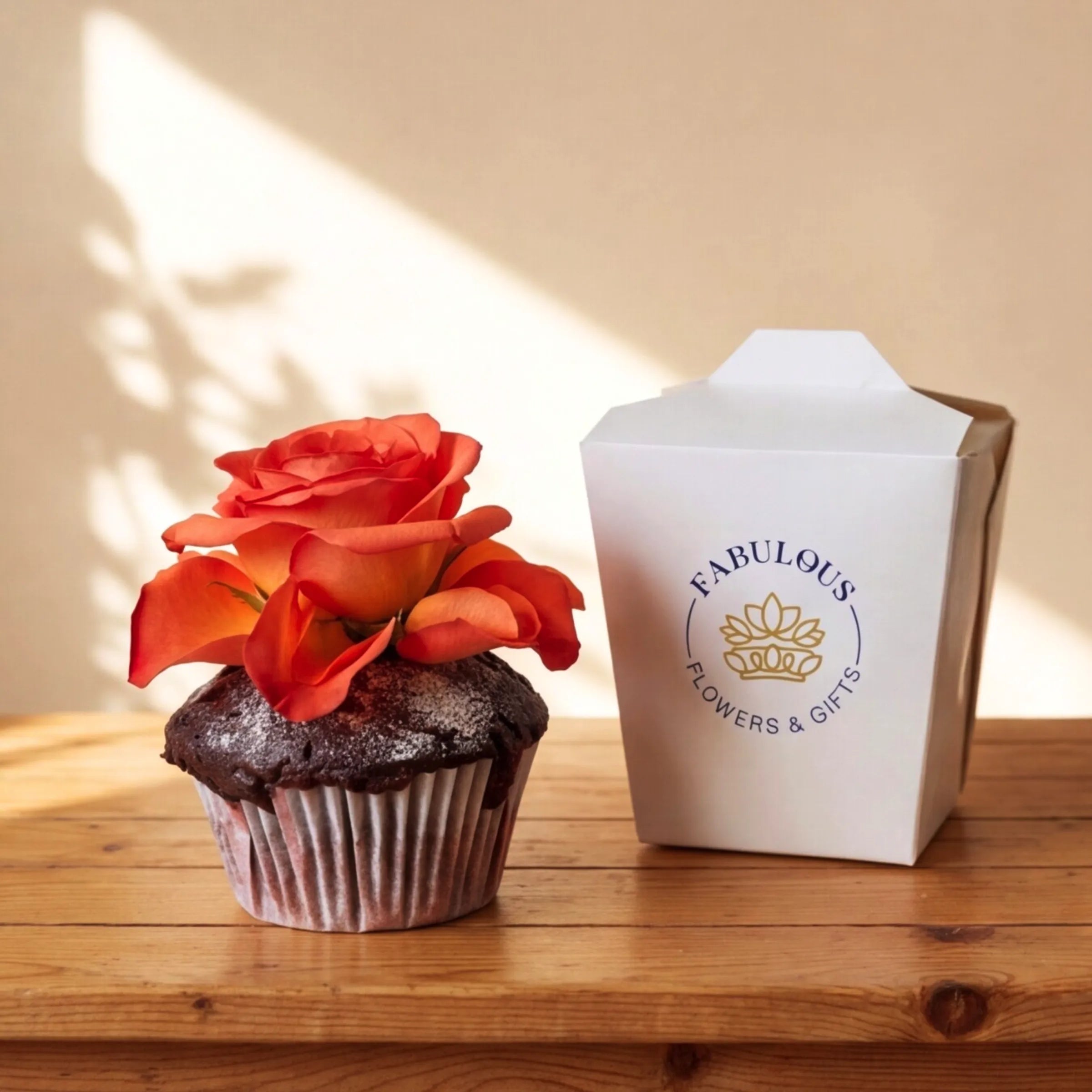 A single chocolate cupcake topped with a vibrant orange rose, sitting beside a white Fabulous Flowers branded gift box with the gold crown emblem, Cape Town same-day cupcake add-on