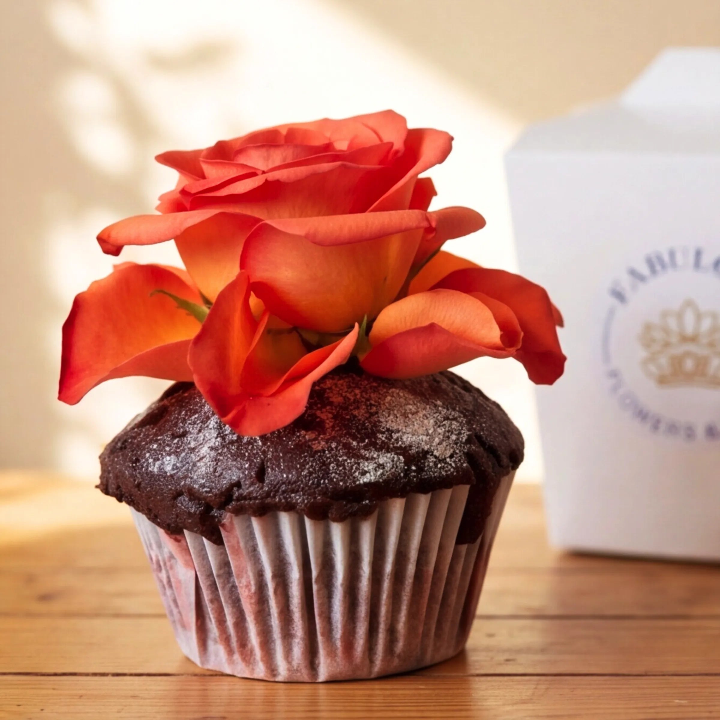 Close-up of a hand-baked chocolate cupcake from Fabulous Flowers with a fresh orange rose topper and a fine icing-sugar dusting on the chocolate dome