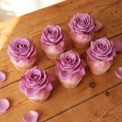 Six vanilla cupcakes arranged in two rows on a rustic wooden table, each topped with a full lavender-pink rose and pink edible glitter, scattered rose petals around them