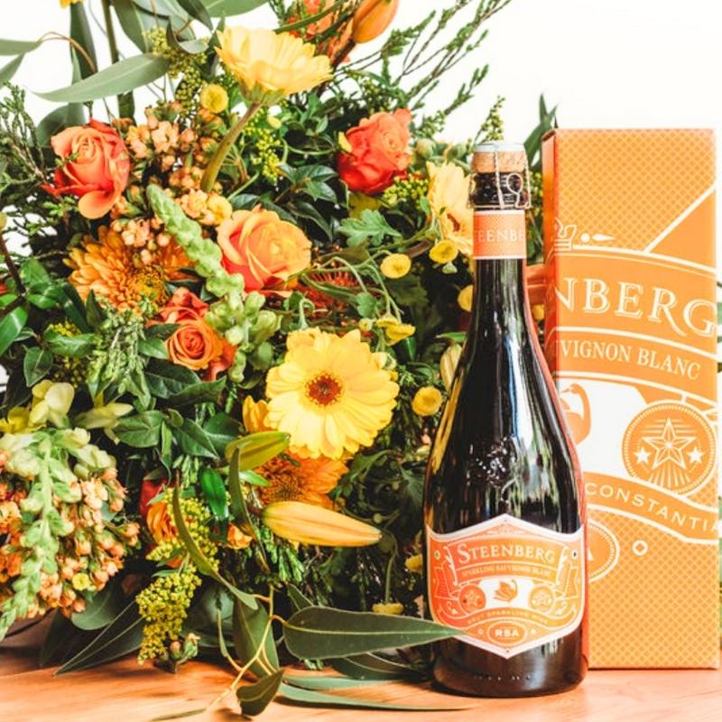 Close-up of Sparkling Orange Bouquet featuring orange roses, gerberas, lilies and lush greenery with celebration bottle