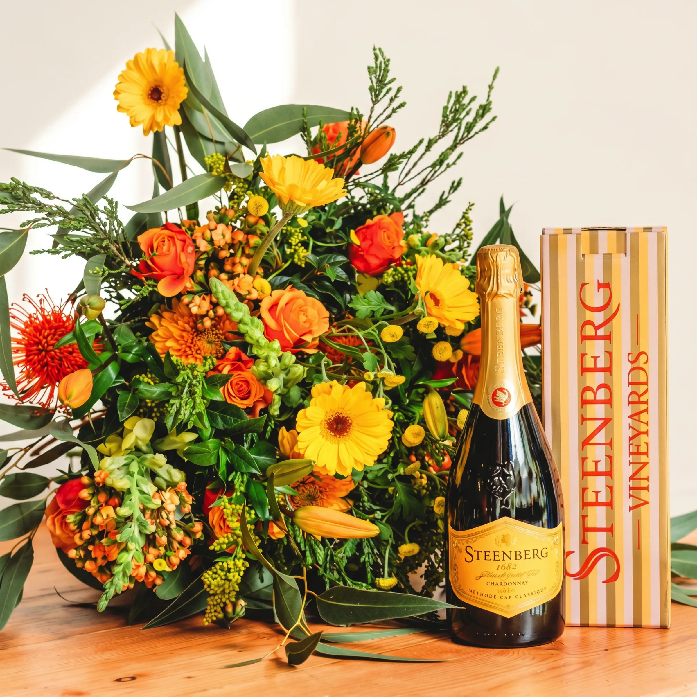 Sparkling Orange Bouquet styled with a bottle of Steenberg 1682 Chardonnay MCC and branded gift box, showing orange roses, yellow gerbera daisies, orange pincushion proteas, yellow craspedia billy buttons, orange tulips, green snapdragons and trailing eucalyptus, Fabulous Flowers South Africa flower and sparkling wine gift