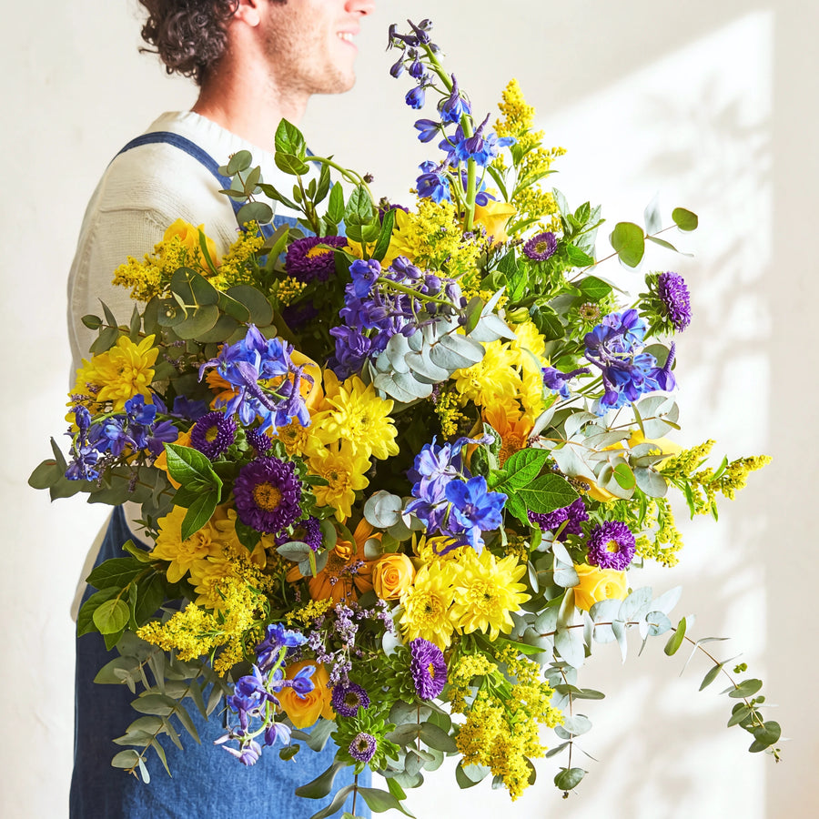Florist holding the Spring Personified spring bouquet of blue delphinium, yellow chrysanthemums and purple asters.