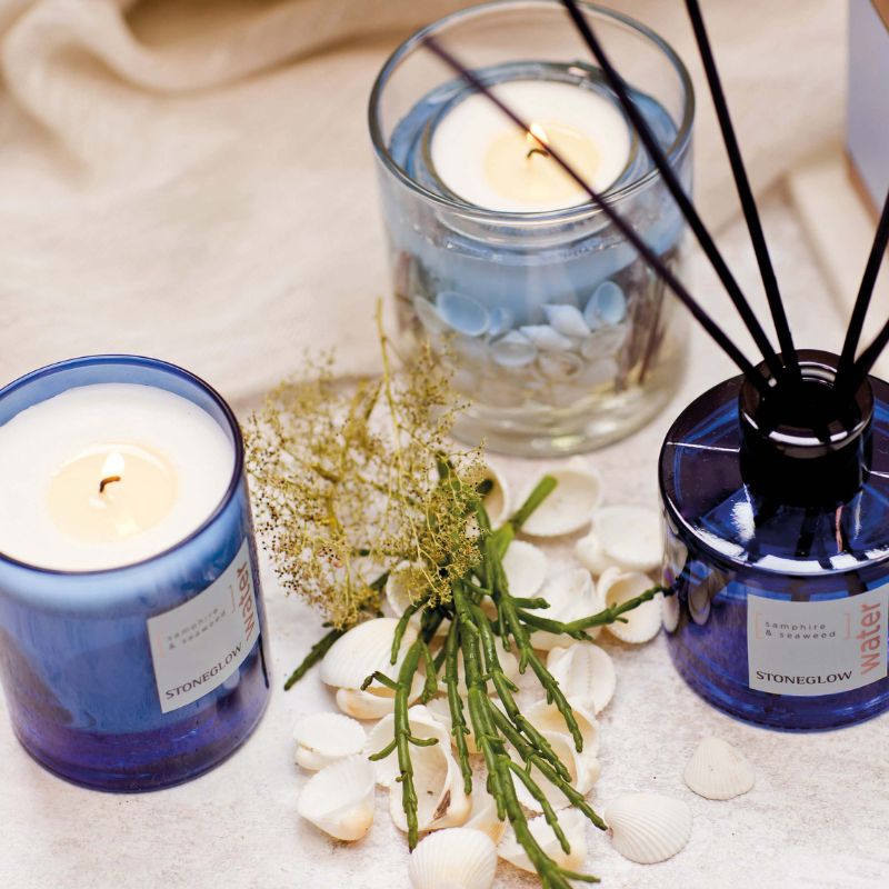 Two blue candles and a diffuser on a textured surface with pebbles and greenery.