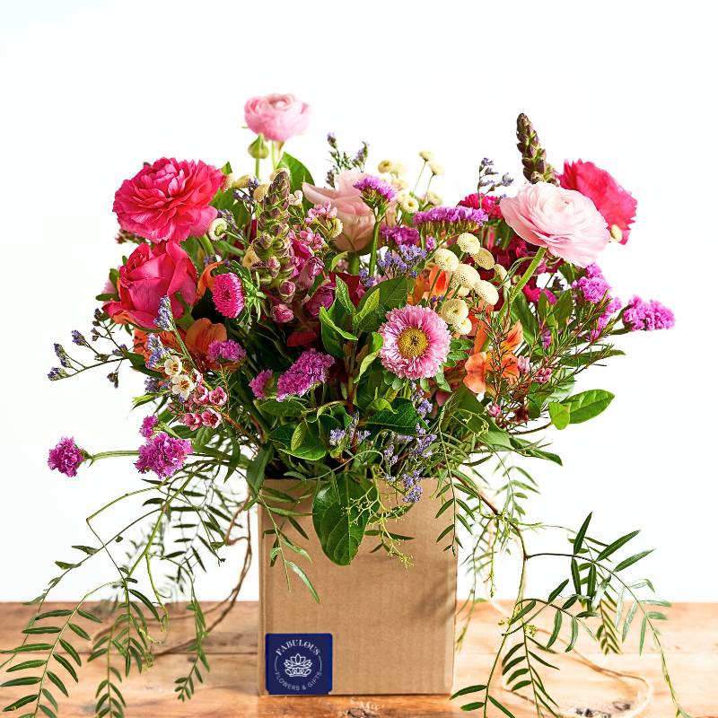 Posy box of colourful flowers in a brown paper bag on a wooden surface with a white background