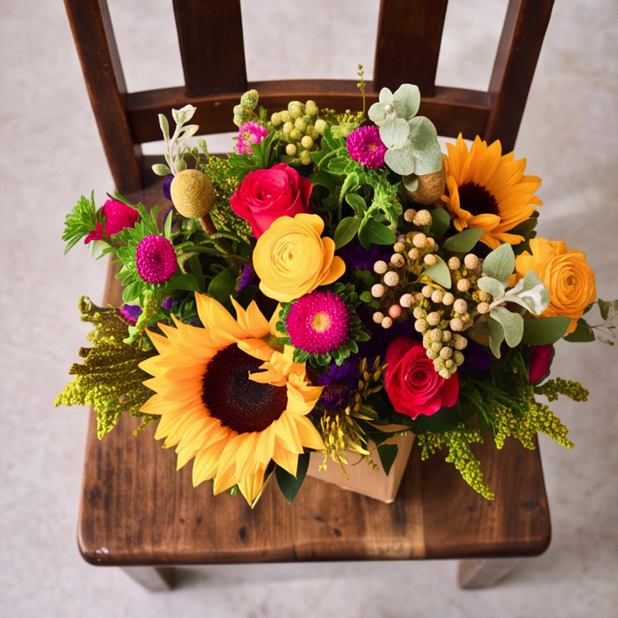 A vibrant artificial flower arrangement featuring sunflowers, roses, and other colorful blooms, displayed on a rustic wooden chair.