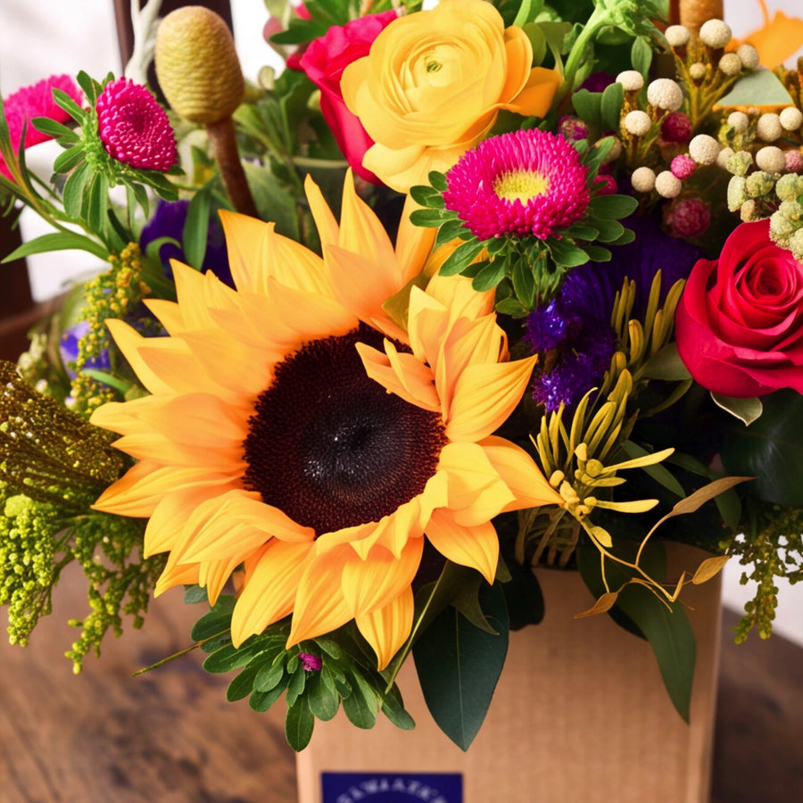Close-up of the Fabulous Flowers Sunflower Posy: a golden sunflower with hot pink roses, yellow ranunculus, magenta asters, purple statice and green craspedia