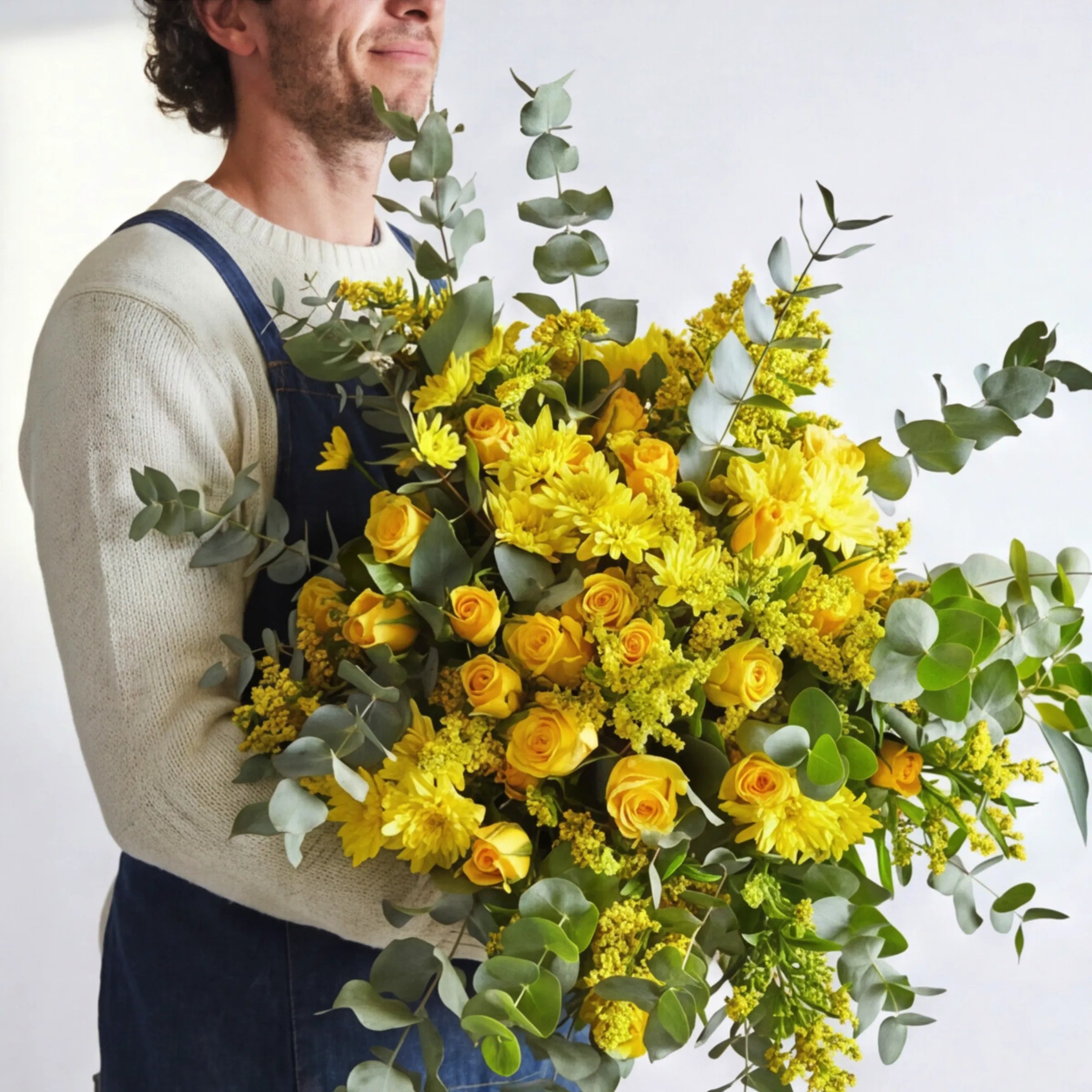 Sunkissed Yellow Bouquet held by a florist in a white sweater and denim overalls, hand-tied arrangement of golden yellow roses, yellow spider chrysanthemums, yellow solidago and trailing silver eucalyptus, Fabulous Flowers South Africa friendship and get-well gift