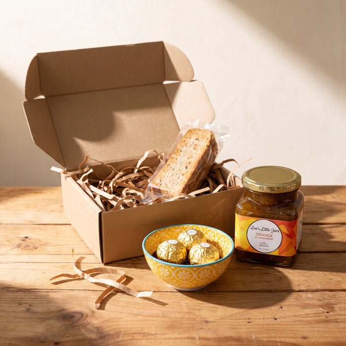 Small kraft gift box with biscotti, beside a yellow daisy-pattern bowl of three Ferrero Rocher and a jar of orange and cardamom jam on a pine table, Fabulous Flowers South Africa