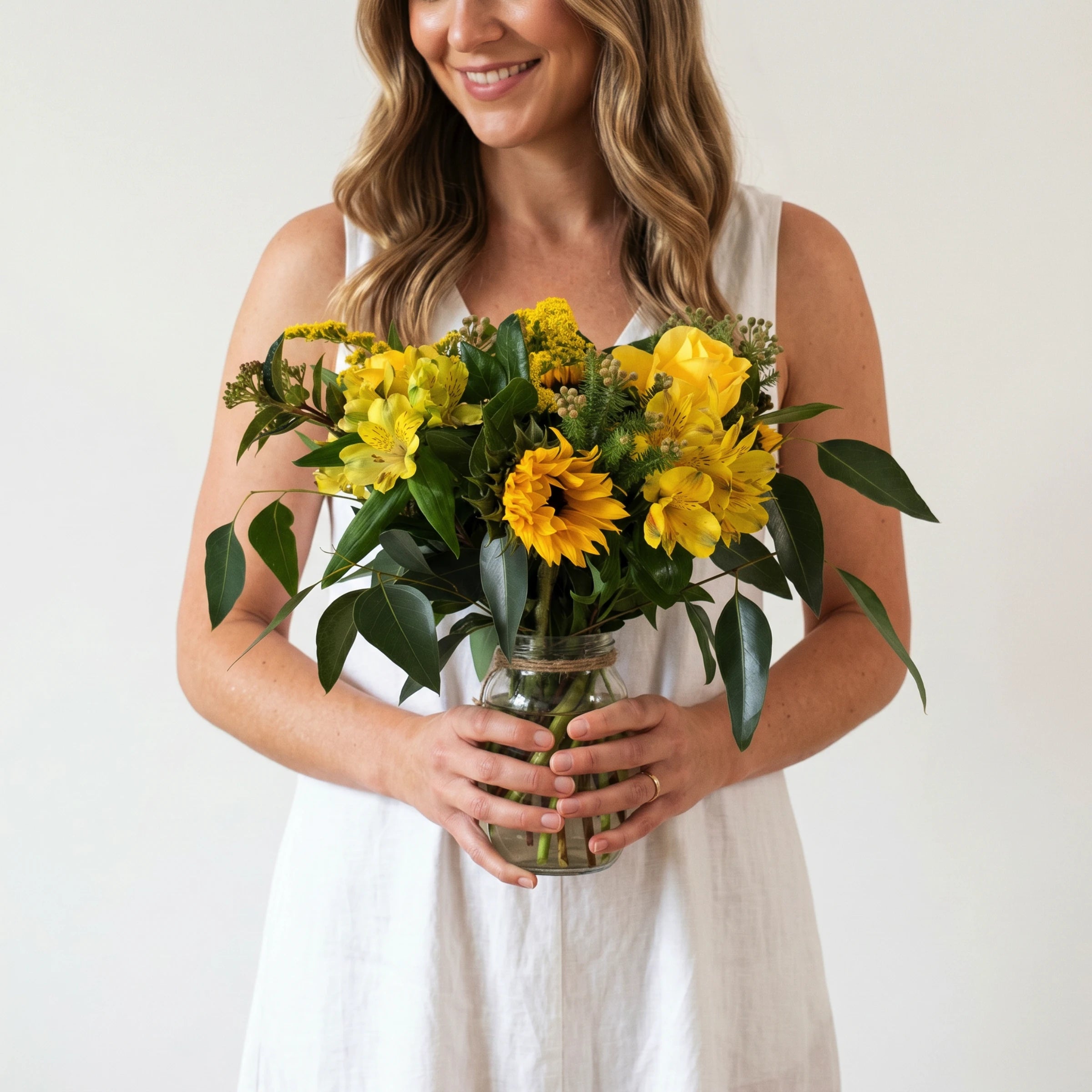 Woman in a white linen dress holding the Sunshine in a Jar arrangement, yellow sunflowers, yellow roses, yellow alstroemeria and solidago with ficus and eucalyptus greenery in a glass mason jar tied with twine, from Fabulous Flowers Cape Town.
