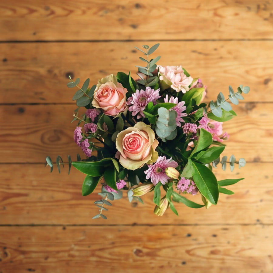 Top view of Sweet Pea in a Jar with peach roses, lilac chrysanthemums, pink statice and silver eucalyptus on a wooden table