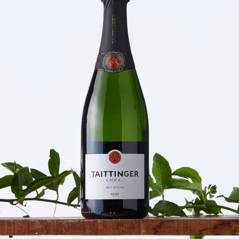 Elegant Taittinger Brut Réserve champagne bottle with black foil and white label, styled with fresh green leaves on a wooden ledge.