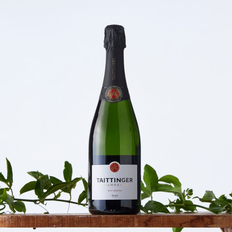Bottle of Taittinger Brut Réserve champagne displayed on a wooden shelf with green vine leaves against a light background.