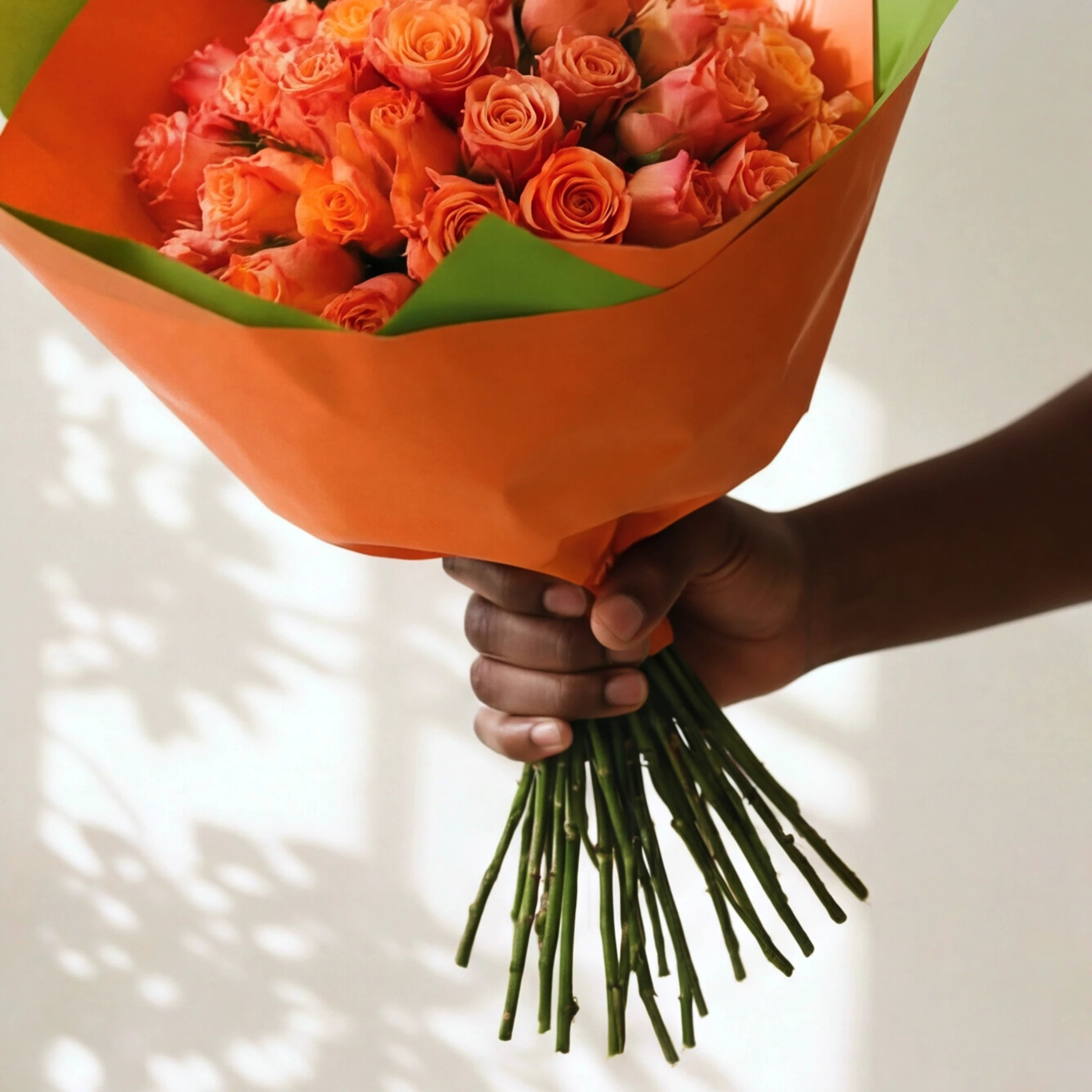 Close-up of a hand holding the orange rose bouquet by its long green stems, roses in orange wrap.