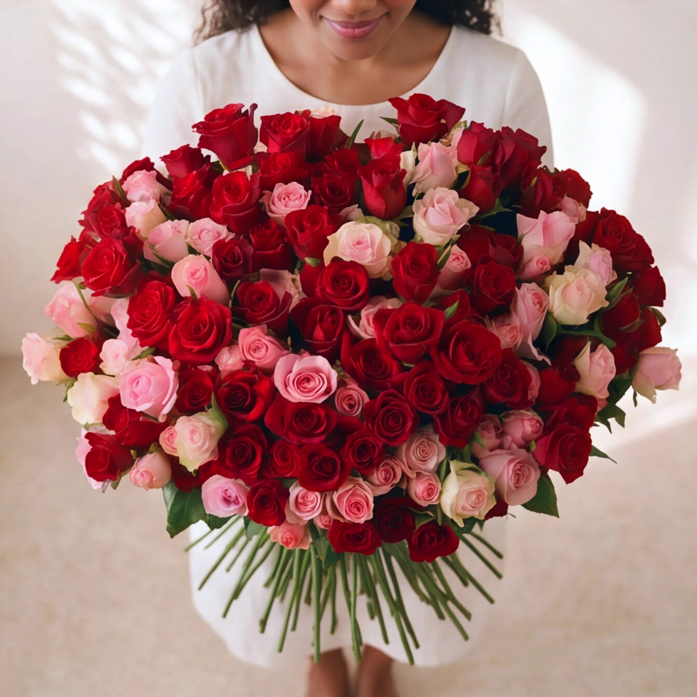 A large, dense bouquet of red and pink roses held by a person wearing a white dress, photographed from above in a bright indoor setting.