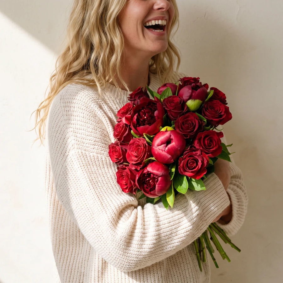 Blonde woman laughing and hugging a bouquet of red peonies and red roses