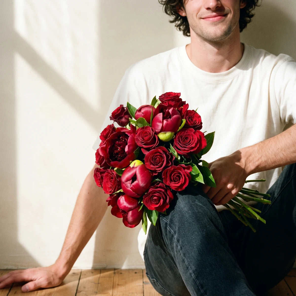 Man sitting against a wall holding a romantic red peony and rose bouquet