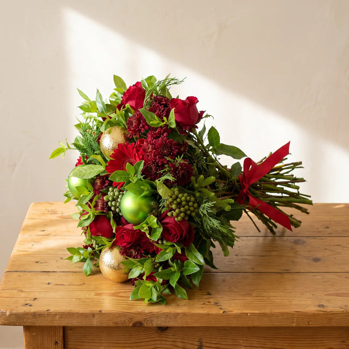 Christmas bouquet of red roses, burgundy chrysanthemums, brunia and gold and lime baubles tied with a red ribbon, lying on a pine table, Fabulous Flowers South Africa