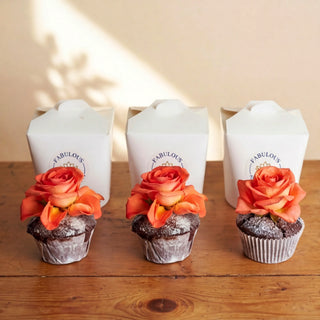 Three chocolate cupcakes lined up on a rustic wooden table in front of three white Fabulous Flowers branded boxes, each topped with a vibrant orange rose and finished with a soft icing-sugar dusting