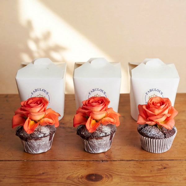 Three chocolate cupcakes lined up on a rustic wooden table in front of three white Fabulous Flowers branded boxes, each topped with a vibrant orange rose and finished with a soft icing-sugar dusting