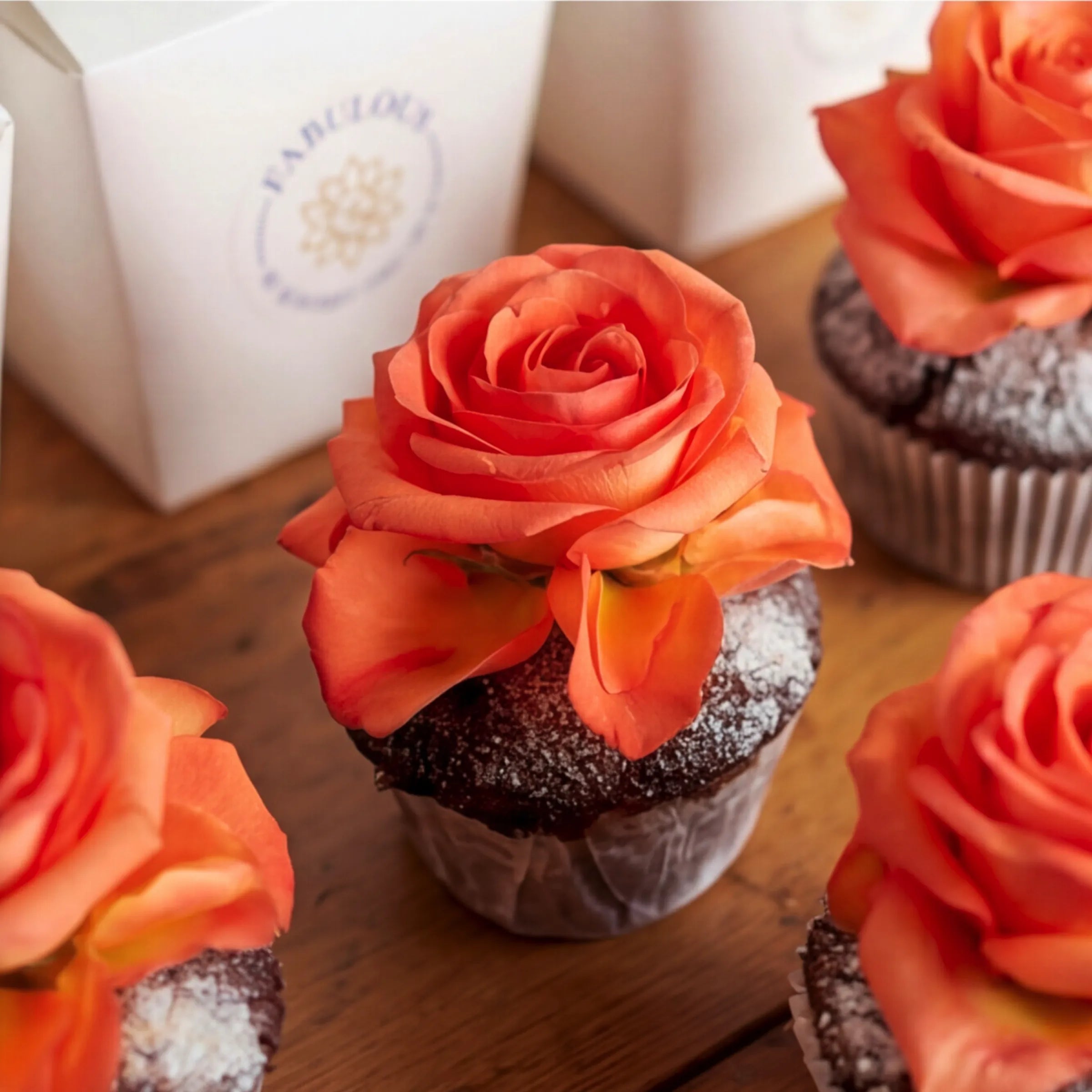 Close-up of Fabulous Flowers chocolate cupcakes with vibrant orange rose toppers and icing-sugar finish on dark chocolate sponge, alongside the white branded gift box