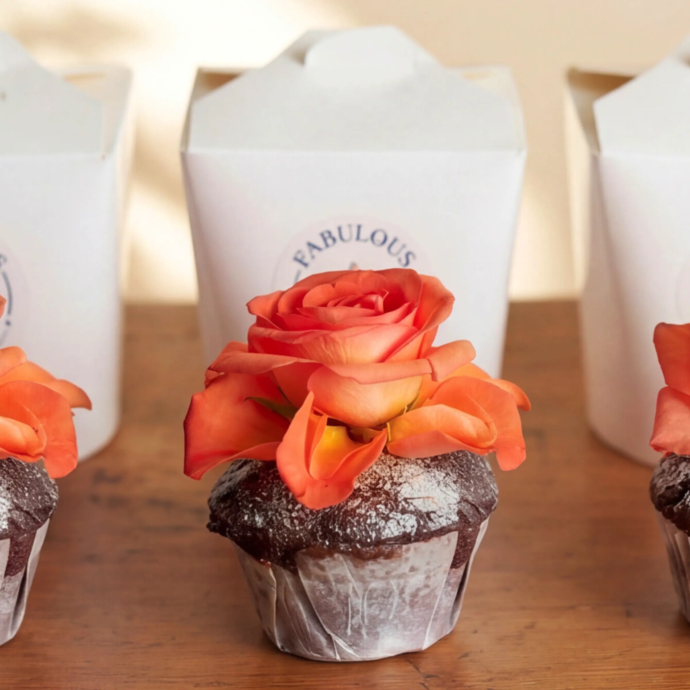 Macro of a single chocolate cupcake from the Three Chocolate Cupcakes set, dark chocolate sponge with icing-sugar dusting and an open orange rose bloom resting on top