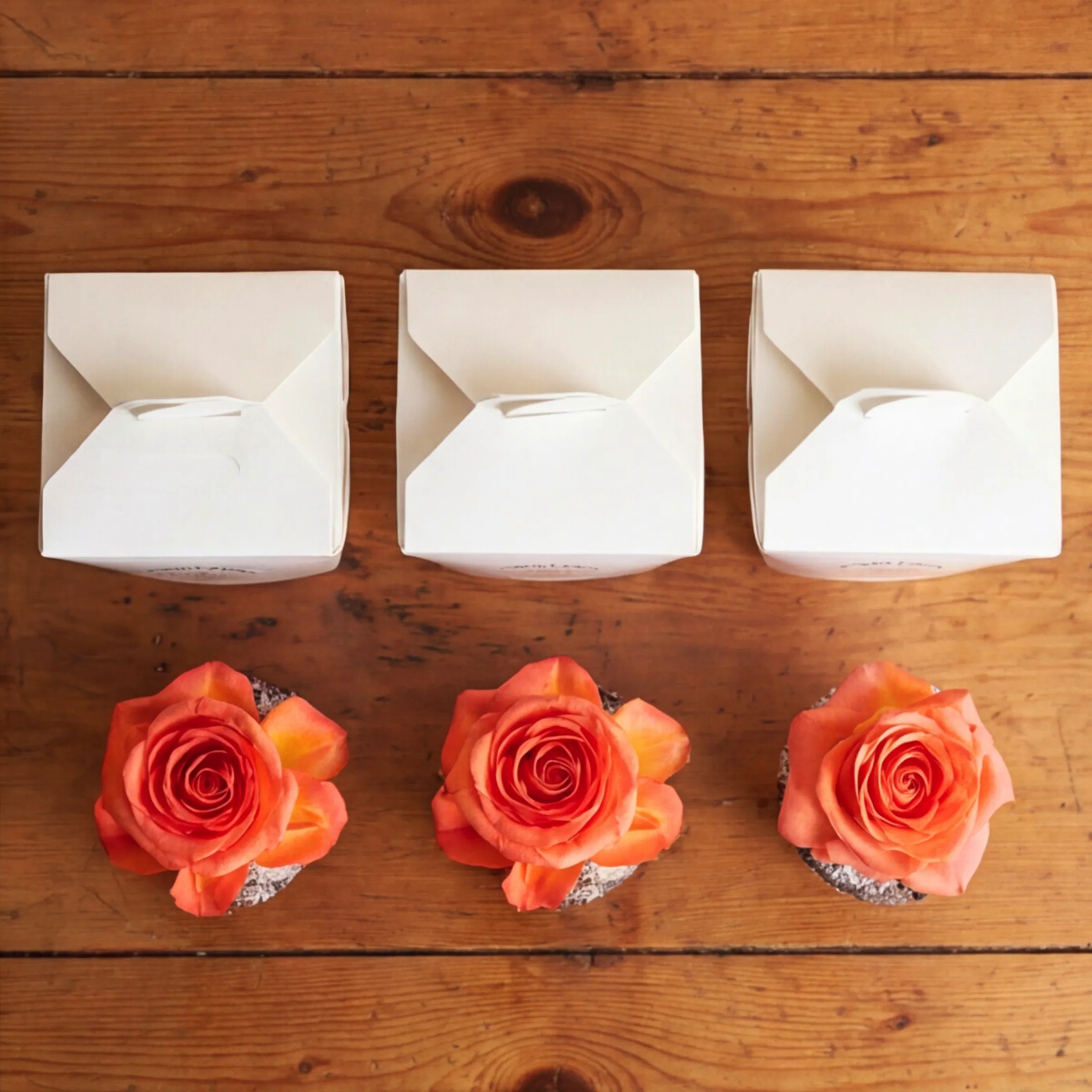 Overhead flat lay of three white Fabulous Flowers branded cupcake boxes paired with three orange rose-topped chocolate cupcakes arranged on a rustic wooden surface