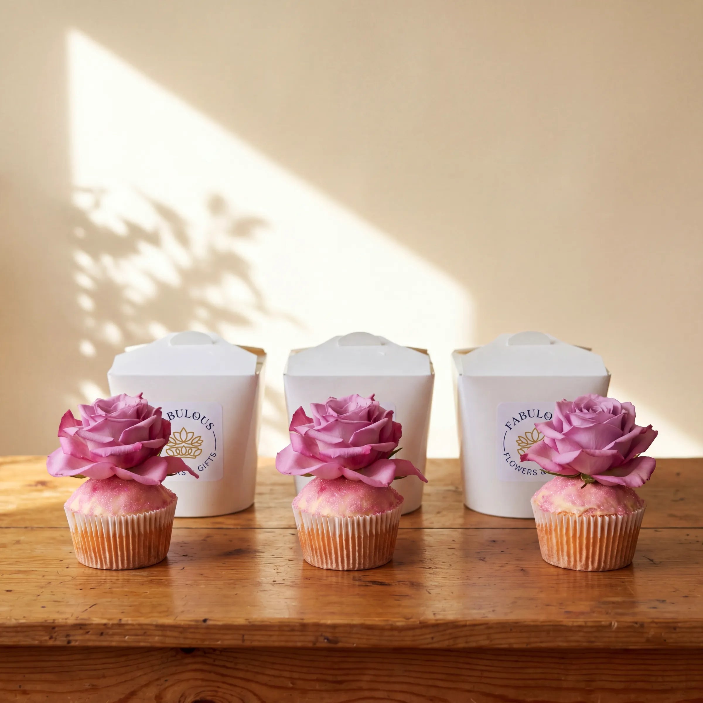 Three vanilla cupcakes lined up on a rustic wooden table in front of three white Fabulous Flowers branded boxes, each cupcake topped with a fresh pink rose and pink edible glitter