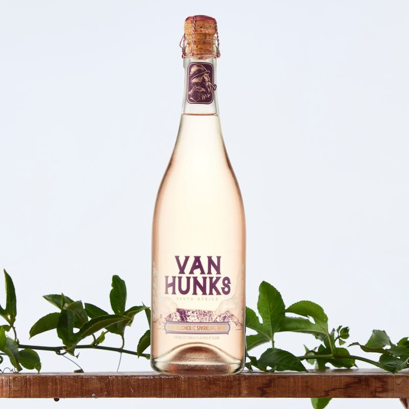 Van Hunks Non-Alcoholic Sparkling Rosé bottle with cork top on wooden shelf, styled with green foliage