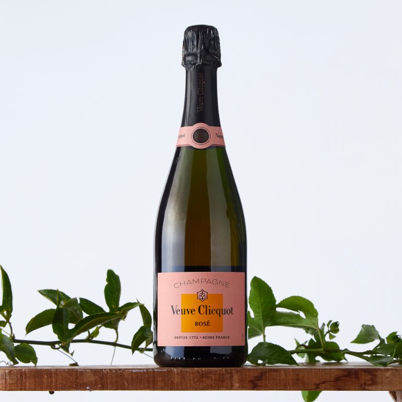 Bottle of Veuve Clicquot Rosé champagne on a wooden shelf with leafy green foliage, featuring the signature pink and orange label.