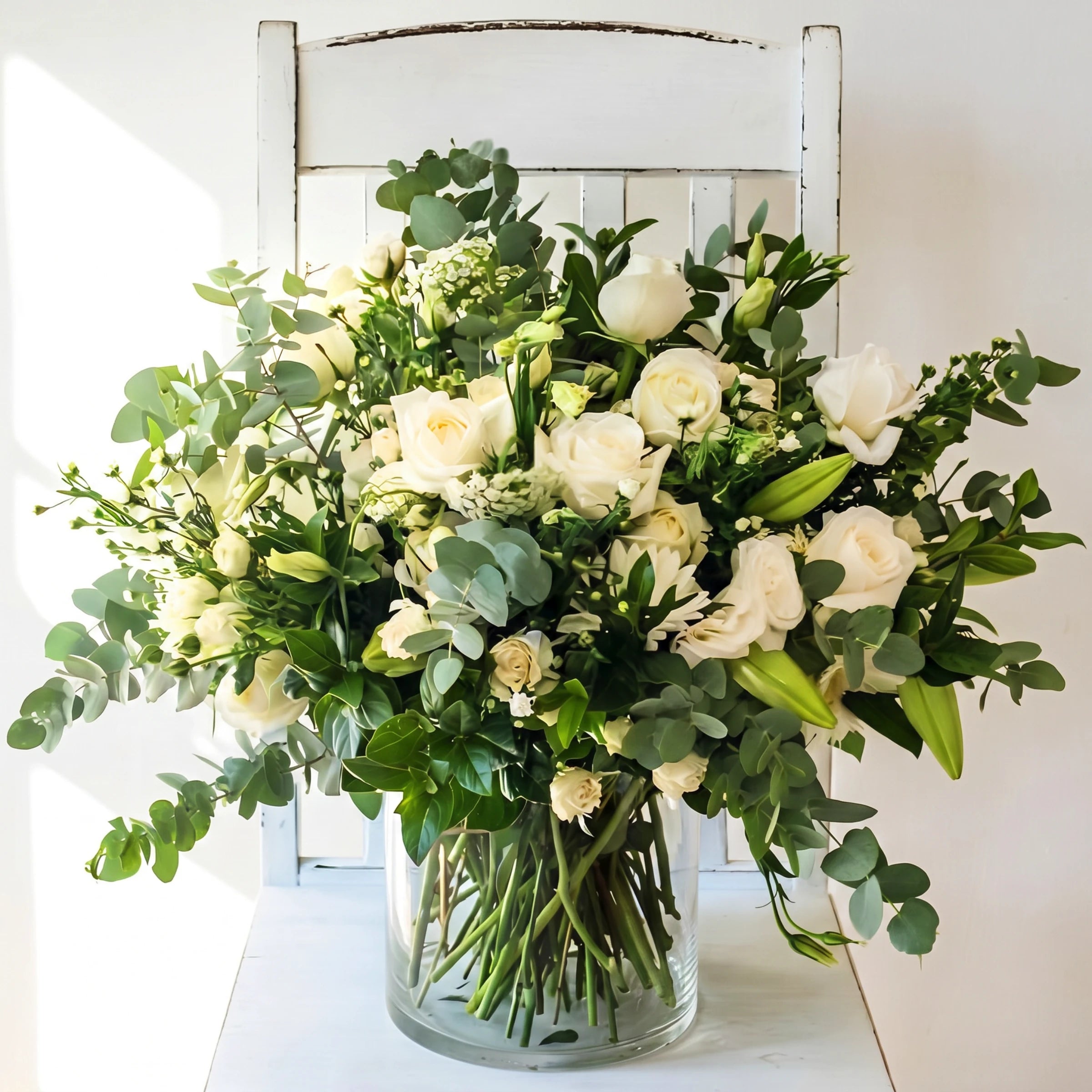 White Purity Vase Arrangement on a rustic white painted chair, white roses, white lisianthus, lily buds, silver dollar eucalyptus and lush greenery in a clear glass cylinder vase from Fabulous Flowers Cape Town