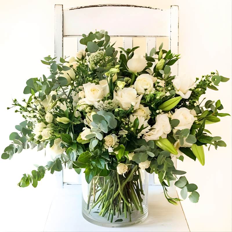 Vase arrangement of white flowers in a clear vase on a white surface with a white chair in the background.