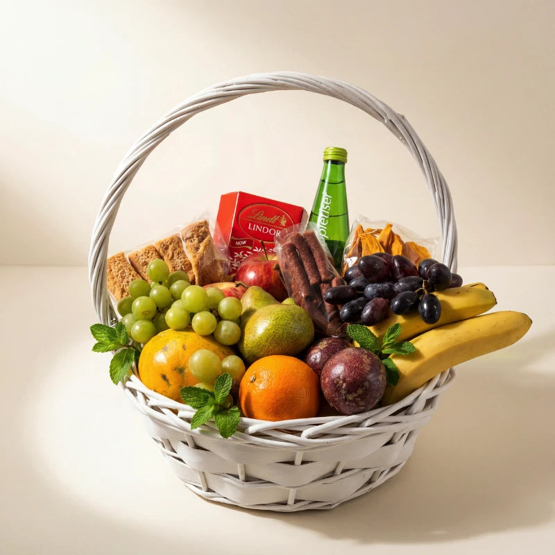 Side view of Wholesome Wishes get well fruit basket with seasonal fruit, Lindt Lindor, Appletiser, biltong and dried mango in a white wicker basket from Fabulous Flowers