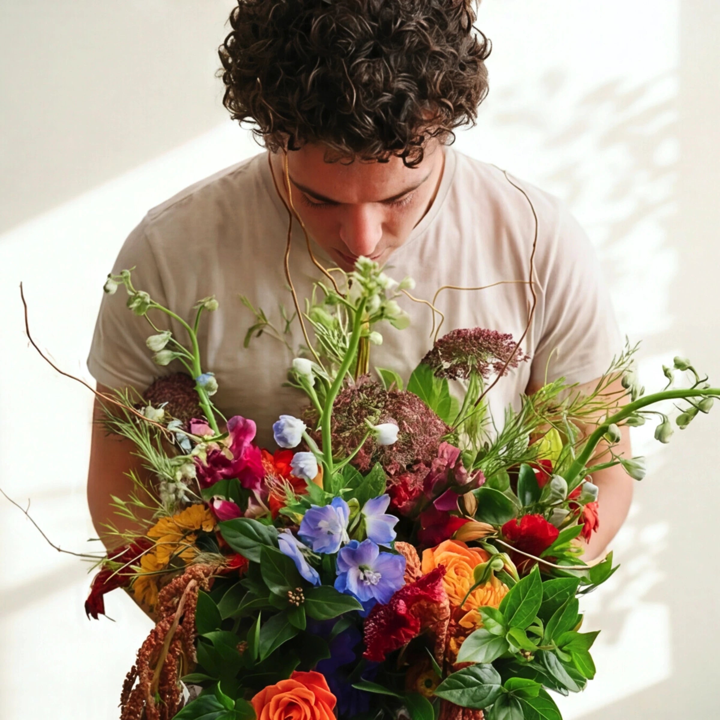 A man with curly dark hair wearing a beige t-shirt leans down to smell a large, vibrant, multicolored wildflower bouquet held in front of him against a bright, sunlit white background.