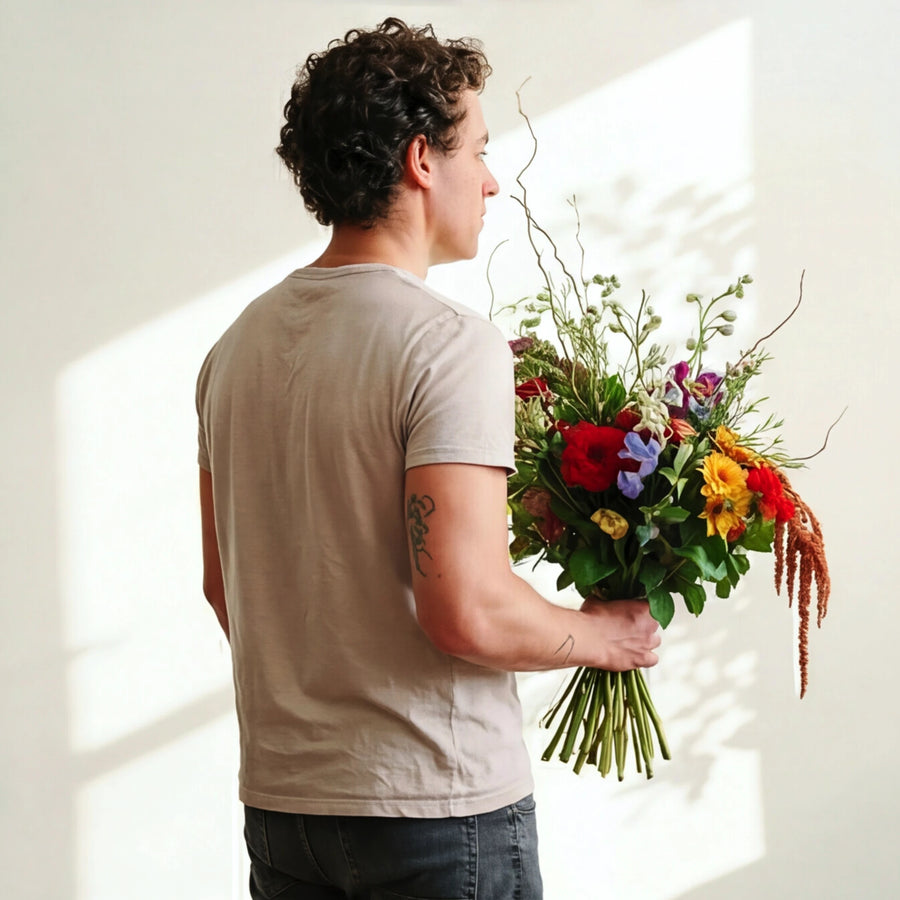 A man in a beige t-shirt and dark jeans stands with his back to the camera, holding a large, vibrant, multicolored flower bouquet with green stems against a warm, sunlit cream wall.