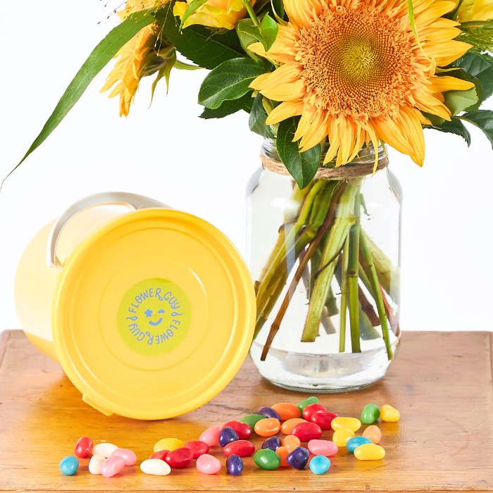 Yellow container with a smiley face logo next to a glass jar with sunflowers and colorful candies on a wooden surface.