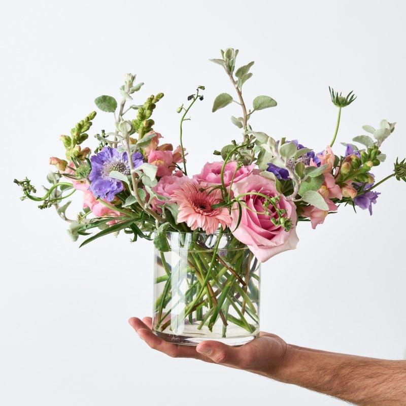 Hand holding a clear glass vase with a bouquet of flowers against a white background