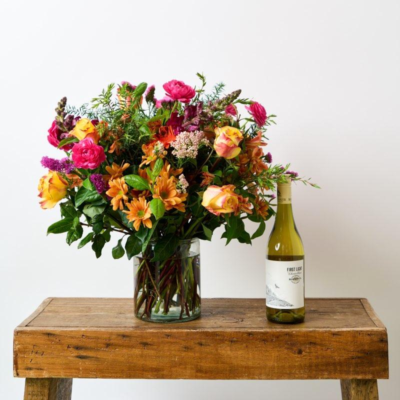 First Spring Blooms flower arrangement is created in a glass case using bright pink, orange and maroon hues and is paired with Remhoogte First Light wine wine from Fabulous Flowers and Gifts