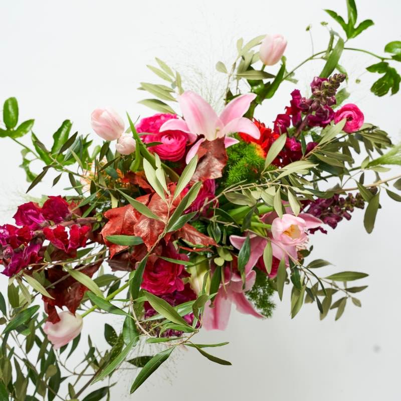 Close up of pastel pink and maroon modern flower arrangement with lilies, ranunculus and whimsical greenery 